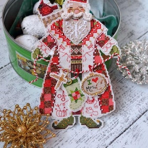 Puede incluir: Un adorno de Santa Claus en punto de cruz con un abrigo rojo y blanco de cuadros, barba blanca y gafas doradas. La figura sostiene un pequeño calcetín, una cruz y una etiqueta circular con el texto I heart Mrs. Claus.