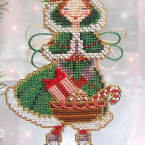May include: A cross-stitch ornament of a figure wearing a green hooded cloak with white trim, holding a brown basket containing a pink gift with a red bow and a candy cane. The figure wears white ice skates and has a red flower on the hood.