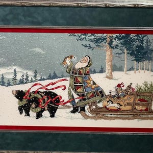 May include: Cross-stitch artwork depicting Santa Claus in a snowy forest scene. Santa, in a patchwork coat, holds a small dog. Two black bears pull a sleigh filled with gifts and greenery. The artwork is framed with a red and green border.
