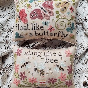 Può includere: Due cuscini rettangolari a punto croce su una base in tessuto color crema. Il cuscino superiore presenta farfalle, viticci e il testo "float like a butterfly". Quello inferiore mostra api, fiori e il testo "sting like a bee". Entrambi hanno bordi intrecciati.