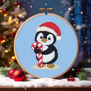 May include: A cross-stitch embroidery hoop featuring a cartoon penguin wearing a Santa hat and holding a candy cane. The penguin is black, white, and brown, set against a light blue background. The wooden hoop is round, and the scene evokes a Christmas theme.