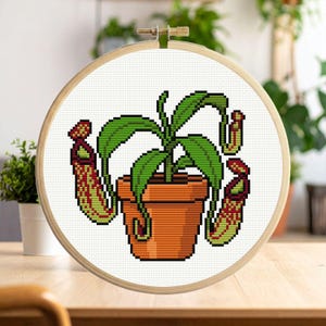 May include: Cross-stitch embroidery of a pitcher plant in a terracotta pot, framed in a wooden hoop. The plant features green leaves and red and green pitcher-shaped traps. The design is on a white fabric background.
