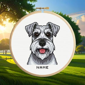 May include: A cross-stitch pattern of a gray and white Schnauzer dog face with a pink tongue, set within a round wooden embroidery hoop. The design features a pixelated style with the word NAME printed in black capital letters below the dog.