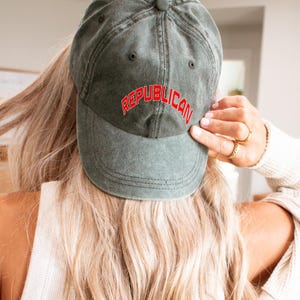 May include: Olive green baseball cap with the word "REPUBLICAN" embroidered in red. The cap is made of a worn, textured fabric. The image shows the back of a person wearing the cap.
