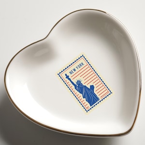May include: A heart-shaped, white ceramic trinket dish with a gold rim. The dish features a blue and red stamp design with the Statue of Liberty and the text "NEW YORK". A decorative item for storing small jewelry or keepsakes.