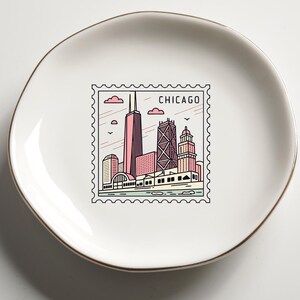 Op de afbeelding: Een witte keramische schaal met een gouden rand. De schaal heeft een Chicago-thema ontwerp dat lijkt op een postzegel, met illustraties van stadsgebouwen in roze, rood en zwart. Het woord "CHICAGO" staat bovenaan de postzegel.