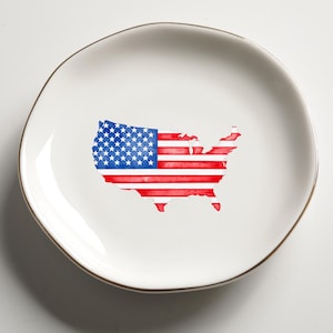 May include: A white ceramic plate with a gold rim. The plate features a watercolor design of the United States map in red, white, and blue, resembling the American flag. The plate has an irregular, organic shape.
