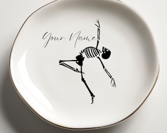 May include: White ceramic plate with a gold rim, featuring a black skeleton illustration in a dancing pose. The plate has the text "Your Name" in a script font. A decorative item for Halloween or themed events.
