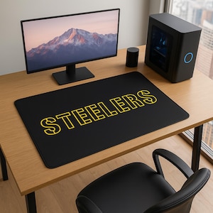 May include: Black rectangular desk mat with the word "STEELERS" in yellow outlined letters. The mat is on a wooden desk with a computer monitor, a computer tower, and a black office chair.