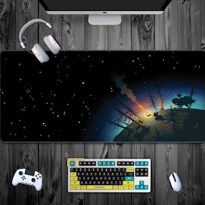 Puede incluir: Una alfombrilla de ratón rectangular grande con una escena de noche estrellada y fogata. La alfombrilla es negra con una ilustración colorida de un planeta, árboles y una fogata. También se ven un teclado, un ratón y un mando blancos.