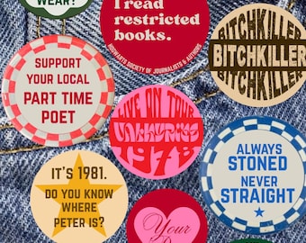 May include: A selection of circular badges with text and illustrations, including "Always Stoned Never Straight" and "I read restricted books." The background is a denim material, with the badges in various colors.