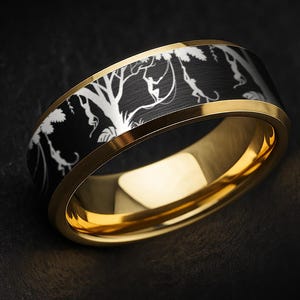 Może przedstawiać: Złoty pierścionek z czarnym paskiem z białym wzorem drzewa. Pierścionek ma polerowane wykończenie i gładki, zaokrąglony profil. Wzór jest inspirowany naturą.
