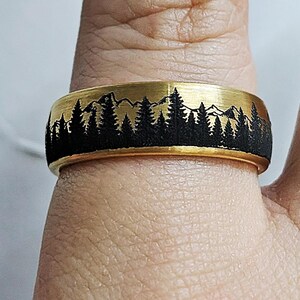 May include: A gold-colored ring with a black forest and mountain design. The ring features a band with a detailed silhouette of trees and peaks. The ring is a piece of jewelry, suitable for everyday wear.