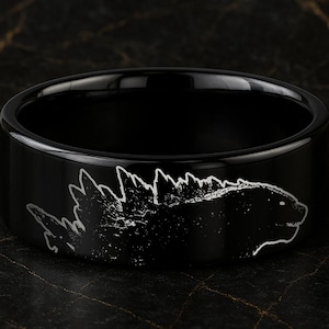 May include: A black ring with a Godzilla design. The ring is black with a silver outline of Godzilla's silhouette. The ring is a wide band style, and the Godzilla design is etched into the band.