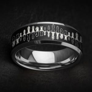 May include: A silver-colored ring with a black band featuring a detailed chess piece design. The ring has a polished, reflective surface and a dark background, highlighting the intricate chess-themed pattern.