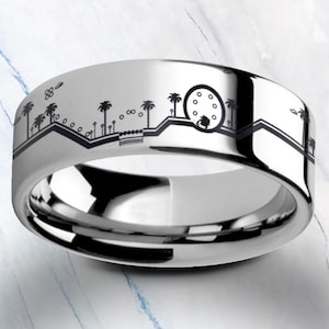May include: A silver ring with a detailed black landscape design. The ring features palm trees, a Ferris wheel, and other elements of a cityscape. The ring has a polished, reflective surface.