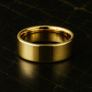 May include: A gold ring with a flat, wide band. The ring's polished surface reflects light, creating a shiny appearance. The design is classic and versatile, suitable for everyday wear or special events.