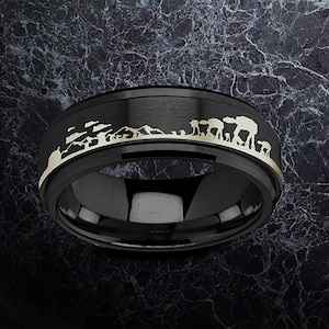 May include: Black ring with a gold-coloured landscape design featuring mountains and a vehicle silhouette. The ring has a polished, reflective surface and a matte black band. A unique piece of jewellery.