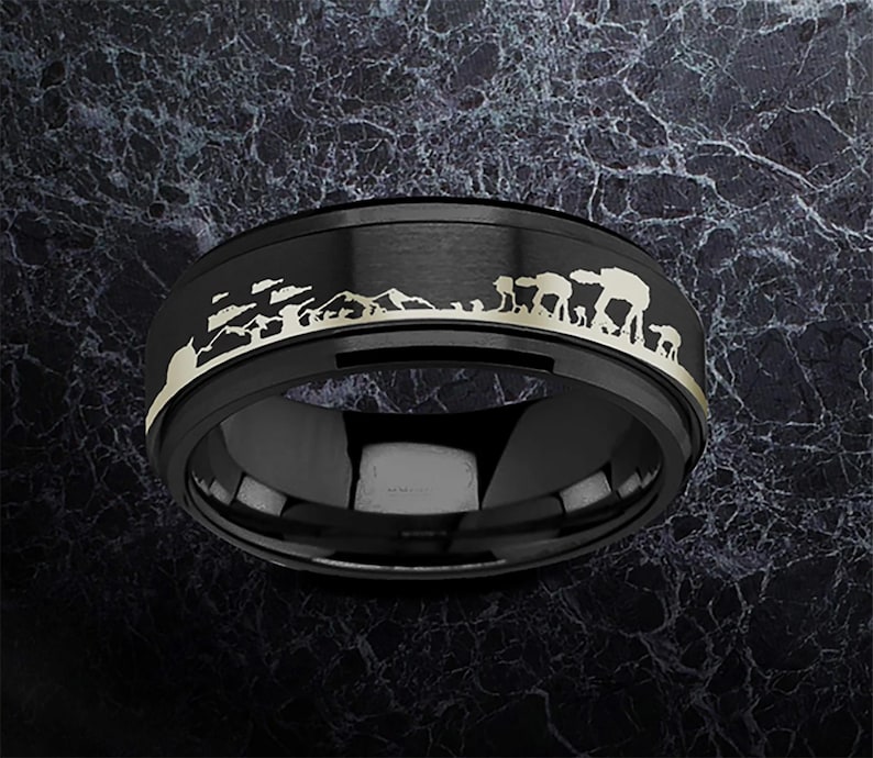 May include: Black ring with a gold-coloured landscape design featuring mountains and a vehicle silhouette. The ring has a polished, reflective surface and a matte black band. A unique piece of jewellery.