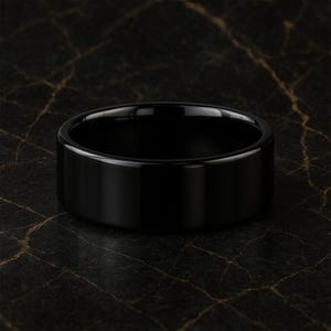 May include: A black, polished ring with a flat, wide band. The ring has a smooth, reflective surface, and the dark colour contrasts with the background. The ring is a piece of jewellery.