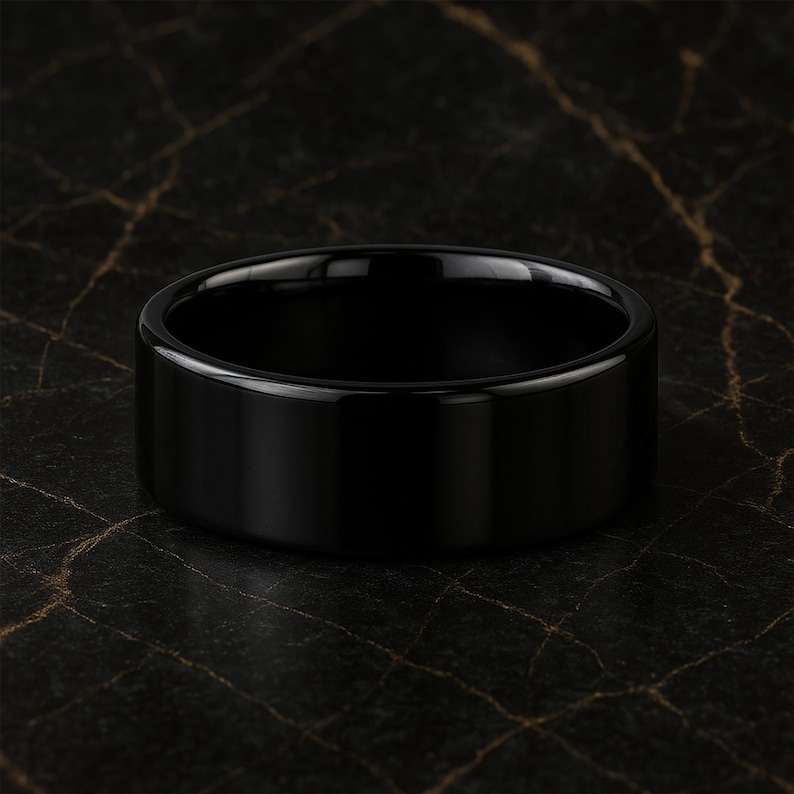 May include: A black, polished ring with a flat, wide band. The ring has a smooth, reflective surface, and the dark colour contrasts with the background. The ring is a piece of jewellery.