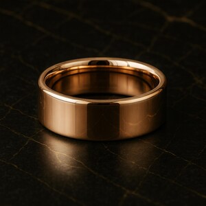 May include: A polished, gold-coloured ring with a flat, wide band. The ring reflects light, showcasing its smooth, metallic surface. The ring's design is simple and modern. The ring's width is approximately 0.8 cm.