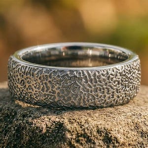 May include: A silver-toned metal ring featuring a highly textured, organic surface pattern that resembles a network of fine, interconnected ridges and valleys. The band has a smooth, polished interior edge and a wide, flat profile with a matte finish.