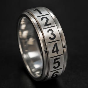 May include: A silver-colored ring with black numerals from 1 to 6. The ring has a rotating outer band with numbers and arrows. The ring is set against a black background.