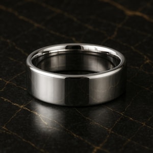 May include: A polished, silver ring with a flat band. The ring has a smooth, reflective surface, and the design is simple and modern. The ring is likely made of stainless steel or a similar metal. The ring is shown on a dark, textured surface.