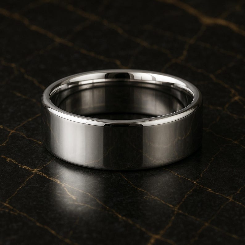 May include: A polished, silver ring with a flat band. The ring has a smooth, reflective surface, and the design is simple and modern. The ring is likely made of stainless steel or a similar metal. The ring is shown on a dark, textured surface.