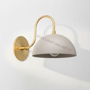 May include: A wall-mounted light fixture with a matte gray dome shade and a gold-colored curved arm. The fixture has a round gold-colored base and a visible light bulb. The text "STAR LIGHT GALLER US" is printed on the shade.