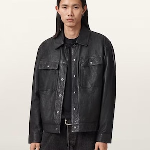 Puede incluir: Chaqueta de cuero negra con dos bolsillos en el pecho y cierres a presión. La chaqueta tiene un cuello clásico y se usa con una camiseta negra y vaqueros oscuros. El modelo lleva un anillo y un pendiente plateados.