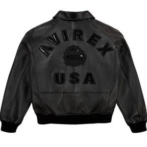 May include: Black leather jacket featuring the word "AVIREX" arched across the back, a circular emblem, and the word "USA". The jacket has a classic bomber style with a ribbed hem and cuffs.