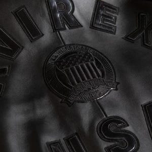 May include: Close-up of a black leather jacket with raised lettering and patches. The jacket features the word "REX" and other lettering. A circular patch with an emblem and the text "All American" and "Established 1975" is also visible.