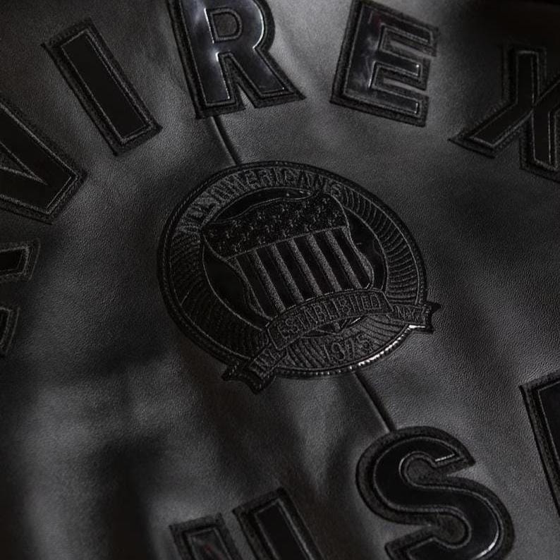 May include: Close-up of a black leather jacket with raised lettering and patches. The jacket features the word "REX" and other lettering. A circular patch with an emblem and the text "All American" and "Established 1975" is also visible.
