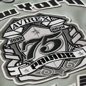 May include: Close-up of a gray leather jacket with embroidered patches. The main patch features the text "AVIREX" and "PALACE" with the number "75" in black and white. Additional patches spell out "NEW YORK".