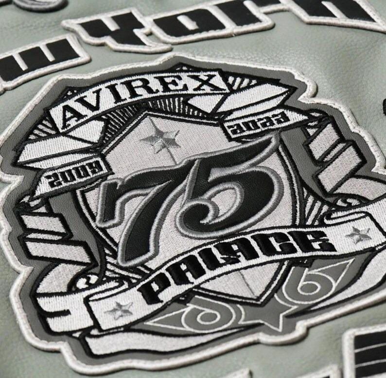 May include: Close-up of a gray leather jacket with embroidered patches. The main patch features the text "AVIREX" and "PALACE" with the number "75" in black and white. Additional patches spell out "NEW YORK".