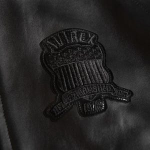May include: Close-up of a black leather jacket with a textured patch. The patch is shield-shaped, with the word "AVIREX" at the top and "ESTABLISHED 1975" on a banner below. The jacket has a subtle sheen.