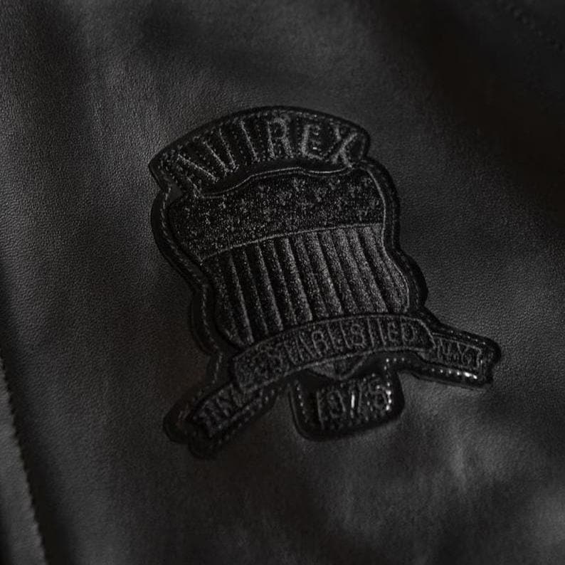 May include: Close-up of a black leather jacket with a textured patch. The patch is shield-shaped, with the word "AVIREX" at the top and "ESTABLISHED 1975" on a banner below. The jacket has a subtle sheen.