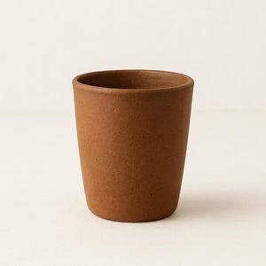 May include: A brown ceramic cup with a slightly textured surface. The cup has a cylindrical shape with a wider opening and a rounded base. The cup is empty and sits on a white surface.