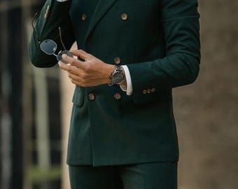 May include: A dark green double-breasted suit with gold buttons. The suit is worn with a white shirt and a black watch. The person is holding sunglasses.