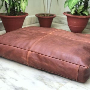 Puede incluir: Un cojín de suelo grande y rectangular de cuero marrón. El cojín tiene un diseño cosido y está colocado sobre una superficie de mármol. Hay plantas en macetas en el fondo.