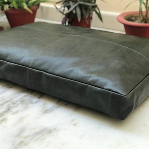 Puede incluir: Un cojín grande y rectangular de cuero verde oscuro. El cojín tiene un diseño cosido y está colocado sobre una superficie de mármol. El cojín es una cama para mascotas.