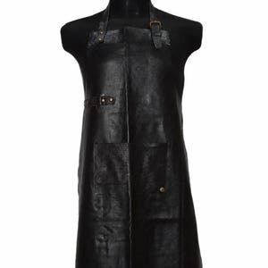 May include: A black leather apron featuring an adjustable neck strap with a metal buckle, a side utility loop with metal rivets, and a large front pocket secured with a single metal rivet. The apron has a textured surface and a smooth finish.