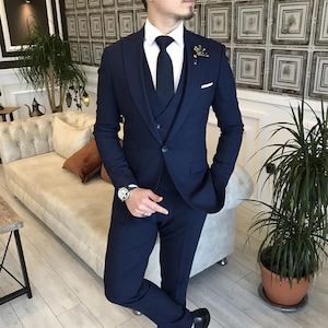 May include: A navy blue three-piece suit with a white dress shirt and black tie. The suit includes a blazer, vest, and trousers. A gold lapel pin and a silver watch are visible. The suit is tailored and appears to be made of a smooth fabric.