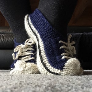 May include: A pair of blue and white knitted slipper shoes with a classic sneaker design. The slippers have white laces and a black trim around the sole.