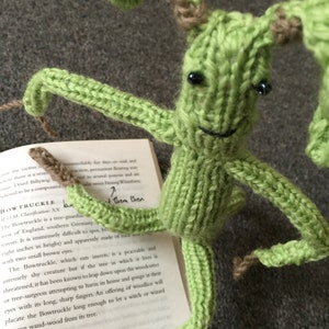 May include: A green knitted toy creature with brown yarn accents and black eyes. The toy is sitting on a book with the text "Bowtruckle" and "Bundimun" visible. The book is about magical creatures.