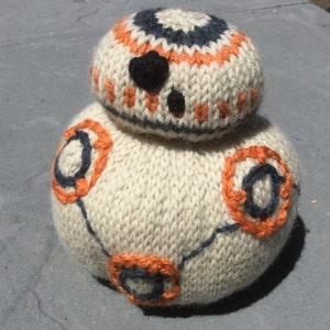 Könnte beinhalten: Ein weiß gestricktes BB-8 Droiden-Spielzeug mit orangefarbenen und schwarzen Details. Der Droide hat einen runden Körper und einen kleineren runden Kopf.