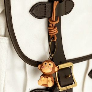 May include: A brown monkey keyring with a leather strap and brass ring. The monkey has a smiling face with pink cheeks and is attached to a white canvas bag with brown leather accents and a brass buckle.