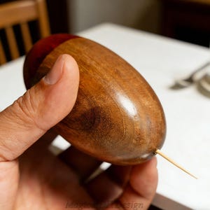Puede incluir: Objeto de madera en forma de bellota con una parte superior de color rojo oscuro y un palillo que se extiende desde la parte inferior. La bellota se sostiene en una mano, mostrando la veta natural de la madera y el acabado liso. El texto "Magical Design" es visible en la parte inferior.